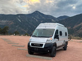Experience mountain adventures! This silver camper van RV rental is parked on a reddish dirt lot, with snow-dusted mountains under a cloudy sky.