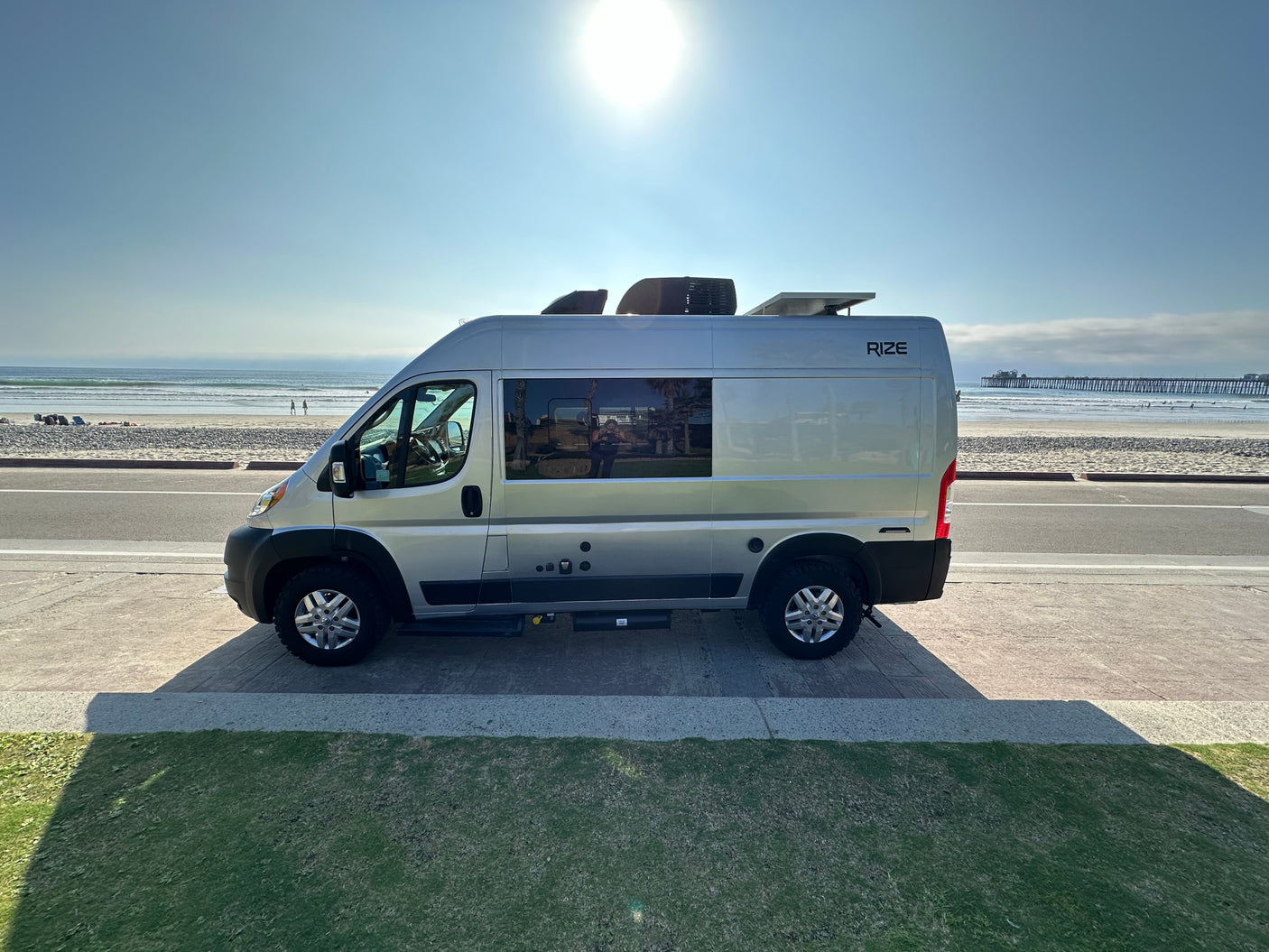 Silver RIZE RV rental parked by a sunny beach with ocean waves and a distant pier, perfect for a coastal adventure.