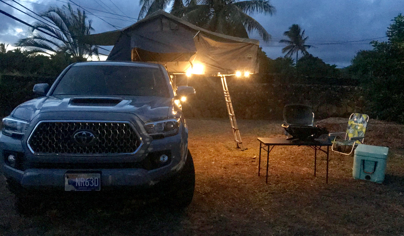 A grey Tacoma truck with a lit rooftop tent & ladder. A grill, table, and cooler are set up for camping. Ready for an RV rental adventure!