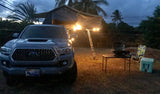 A grey Tacoma truck with a lit rooftop tent & ladder. A grill, table, and cooler are set up for camping. Ready for an RV rental adventure!