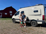 A family with two kids and a dog poses with their white Winnebago Revel RV rental in front of a red barn.