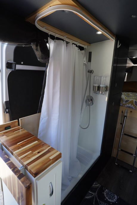 Compact shower in an RV rental, featuring a white curtain, wood counter, soap dispensers, and a glimpse of the bed area.