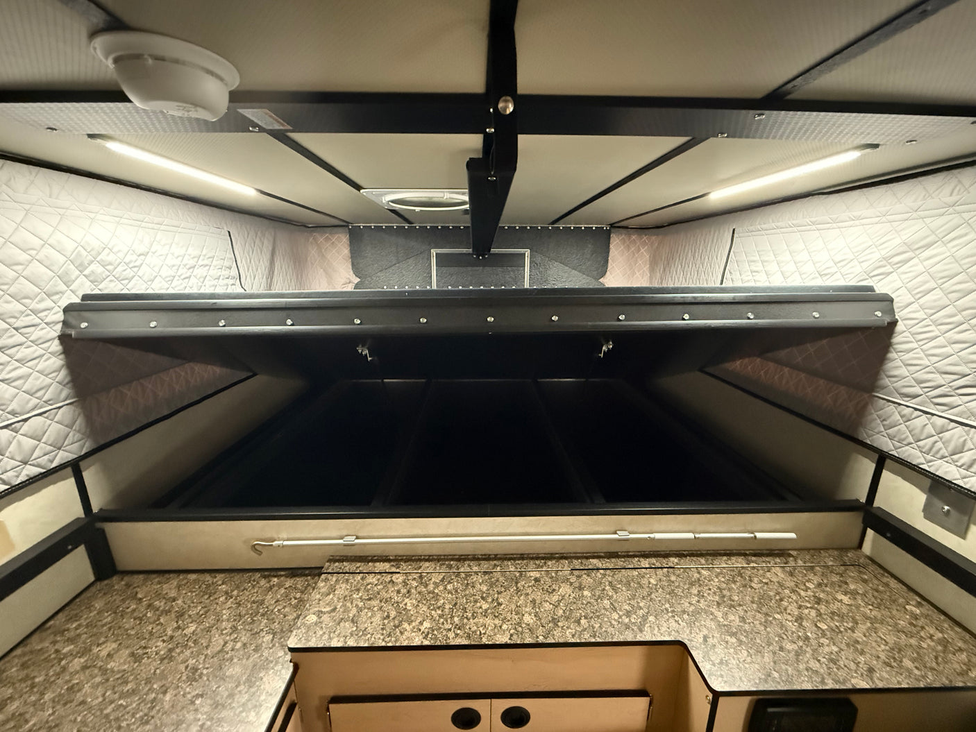 Looking up inside an RV rental pop-up camper: textured ceiling with LED lights, smoke detector, raised bed, and speckled counter.