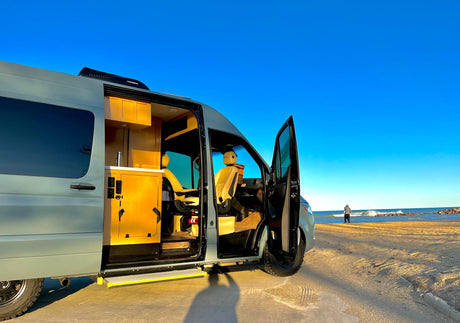 Experience van life! Modern RV rental parked on a beach. Open doors reveal a cozy interior, person by the water.