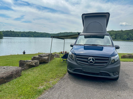 Scenic rv rental: Mercedes pop-top camper van with awning by a tranquil lake, surrounded by green forests, perfect for nature getaways.