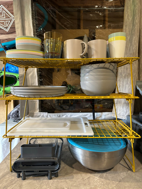 Efficient RV rental kitchen: yellow shelves hold stacked colorful dishes, mugs, and a drying rack with cutting boards.