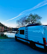 White rv rental parked by a serene lake with a dock, surrounded by lush trees under a blue sky.