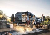 A group of friends enjoys an outdoor grilling experience with their RV rental. Food sizzles over hot coals in a rustic setting.