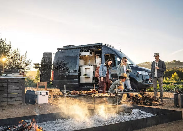 A group of friends enjoys an outdoor grilling experience with their RV rental. Food sizzles over hot coals in a rustic setting.
