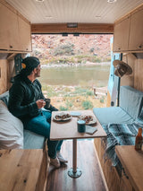 A man enjoys breakfast in a cozy RV rental, gazing at a serene river and rocky mountains from his van.