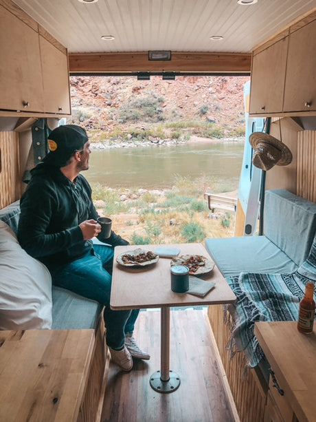 A man enjoys breakfast in a cozy RV rental, gazing at a serene river and rocky mountains from his van.