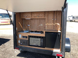 Open hatch of an RV rental revealing a compact mobile kitchen with sink, two-burner stove, microwave, and mini-fridge.