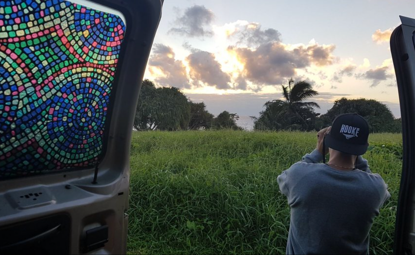 Vibrant mosaic RV rental window frames a person with a HOOKE hat enjoying a picturesque ocean sunset over lush fields.