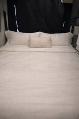 Cozy RV rental bed with white linens & a 'SUN SEA SAND' pillow, set against black window coverings, ideal for travel.