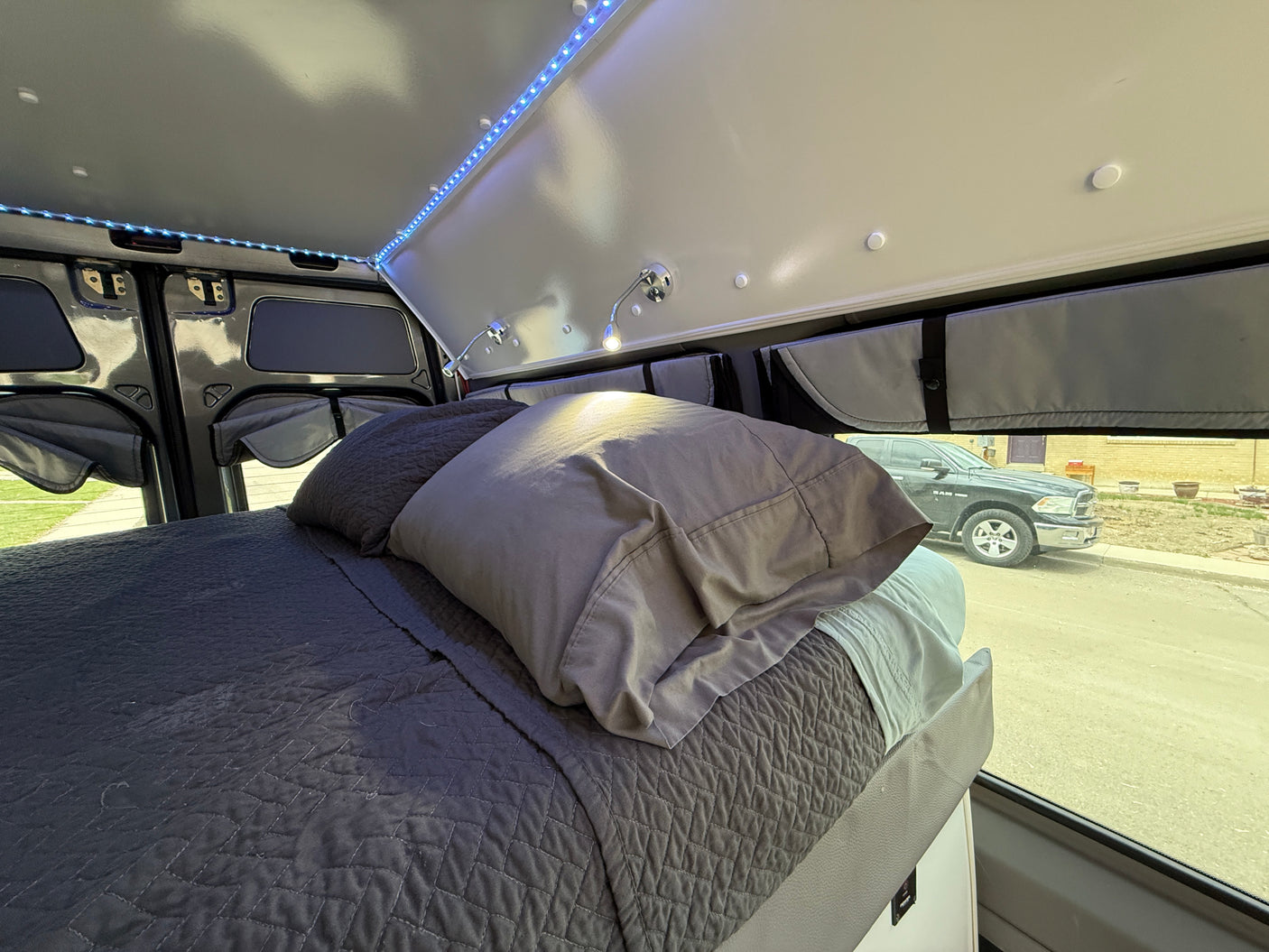 Inside a custom RV rental: a comfy bed with quilted bedding, ambient LED lights, and practical window coverings.