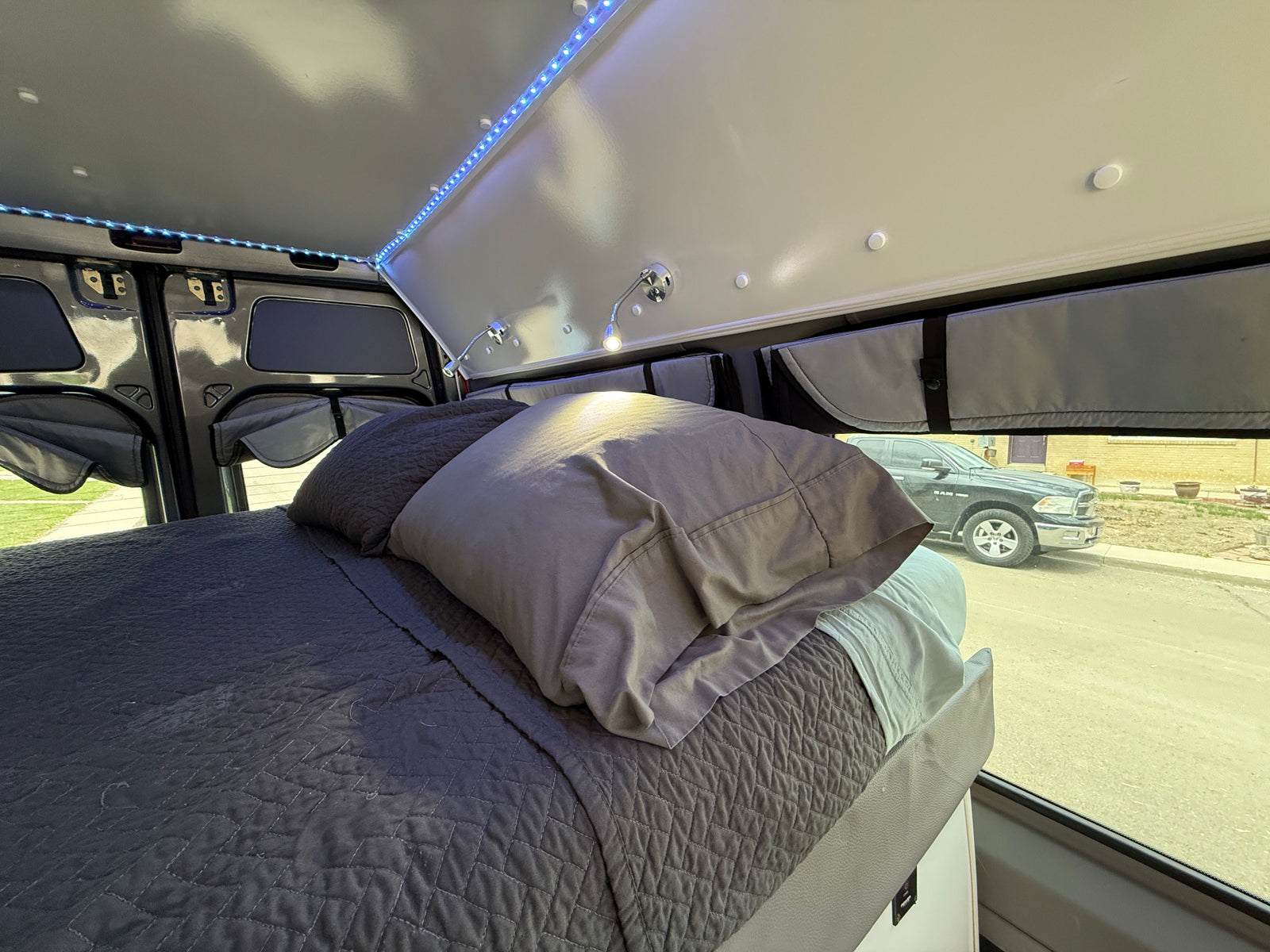 Inside a custom RV rental: a comfy bed with quilted bedding, ambient LED lights, and practical window coverings.