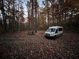 White Ford Transit van (RV rental) parked in a fall forest clearing, covered in leaves. Ideal for camping trips.