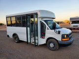 White shuttle bus with open doors on gravel, next to an RV on a trailer under a sunset sky. Potential rv rental.
