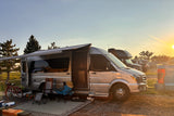 Relaxing by an RV rental camper van at sunset. Chairs, bike, and awning create a cozy campsite scene amidst trees and fields.