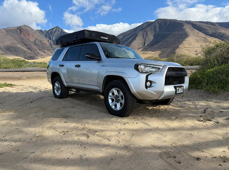 Silver Toyota 4Runner rv rental with a rooftop tent on sand, backed by mountains. Perfect for off-road camping adventures.