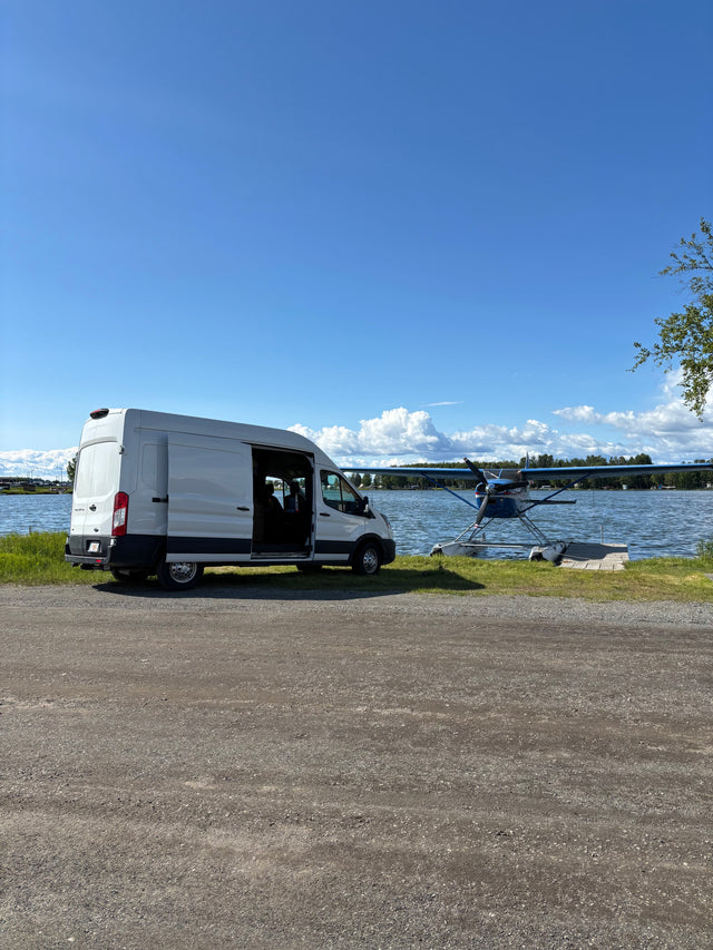 A white RV rental van parked on a gravel shore next to a seaplane on a sunny lake.
