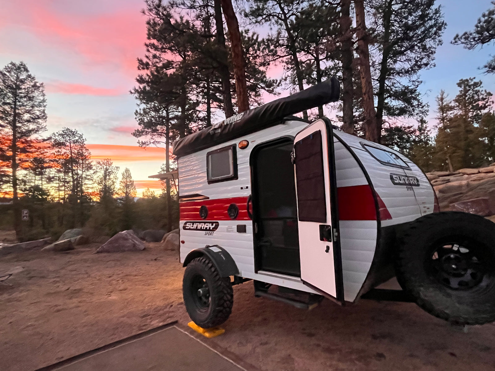 A Sunray Sport teardrop RV rental glows under a stunning pink sunset at a rustic campsite, ready for your outdoor journey.