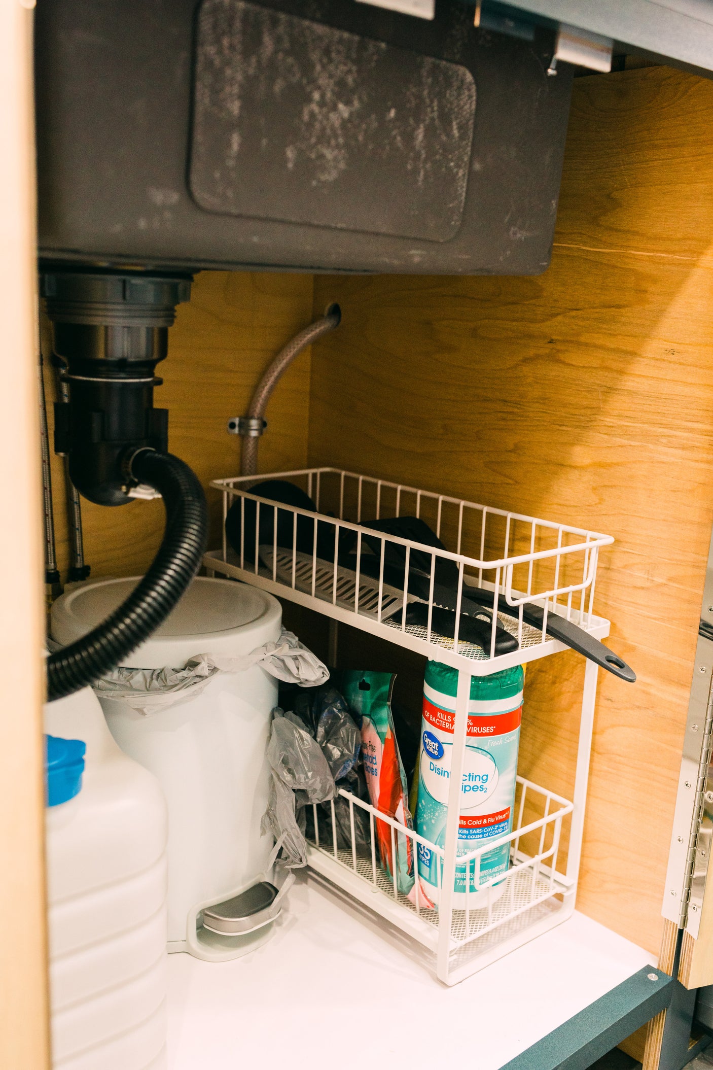 Organized under-sink space in an RV rental, featuring plumbing, a small trash can, and a tiered rack with cleaning supplies.