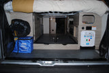 Rear view of an RV rental's organized storage area. Features include a bed overhead, Lynx Levelers bag, and a utility panel for water connections.