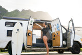 A woman takes an outdoor shower from her custom camper van, a surfboard nearby. Ideal for your next RV rental!