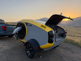 Modern RV rental teardrop camper with open rear, hitched to an electric truck. Scenic sunset over mountains for outdoor travel.