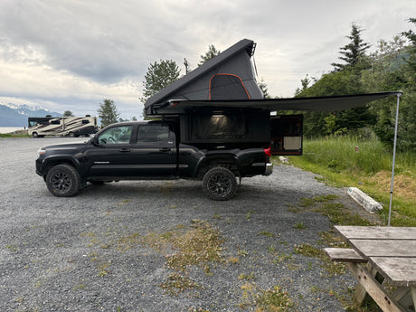 Modified black truck with rooftop tent & awning. An RV rental is visible by mountains & water in a gravel campground.