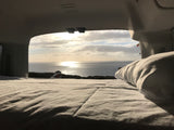 Relaxing with an ocean view from the bed of an RV rental. Sunlight glints on the water; a person watches, and a paraglider flies.