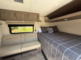 Bright RV rental interior featuring a cozy bedroom area with plaid bedding, comfortable seating, window views, & ample storage.