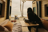 Serene beach view from an RV rental's open door. A woman watches a pedal bike go by, cameras ready for coastal memories.