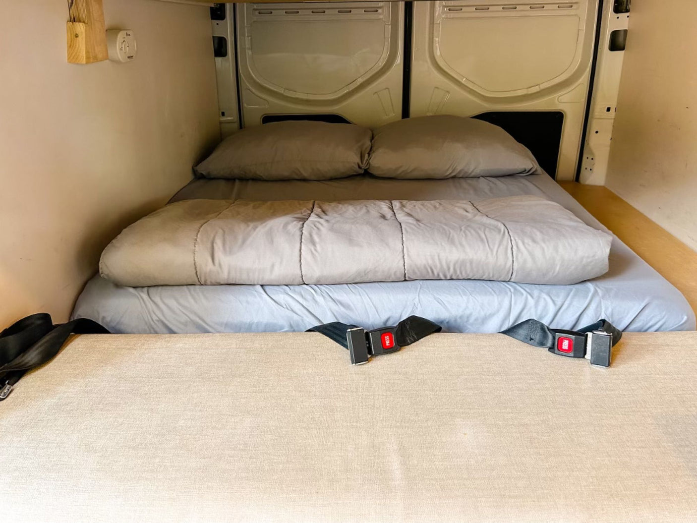 This RV rental features a cozy bed with gray linens, seatbelts, and paneling, ideal for van adventures.