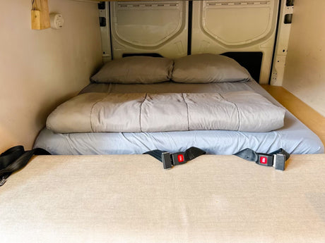 This RV rental features a cozy bed with gray linens, seatbelts, and paneling, ideal for van adventures.