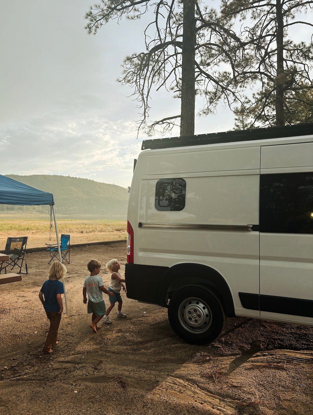 Three kids near a white RV rental camper van at a scenic campsite. Perfect for family outdoor adventures!