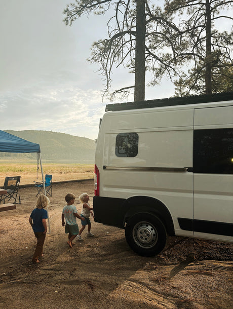 Three kids near a white RV rental camper van at a scenic campsite. Perfect for family outdoor adventures!
