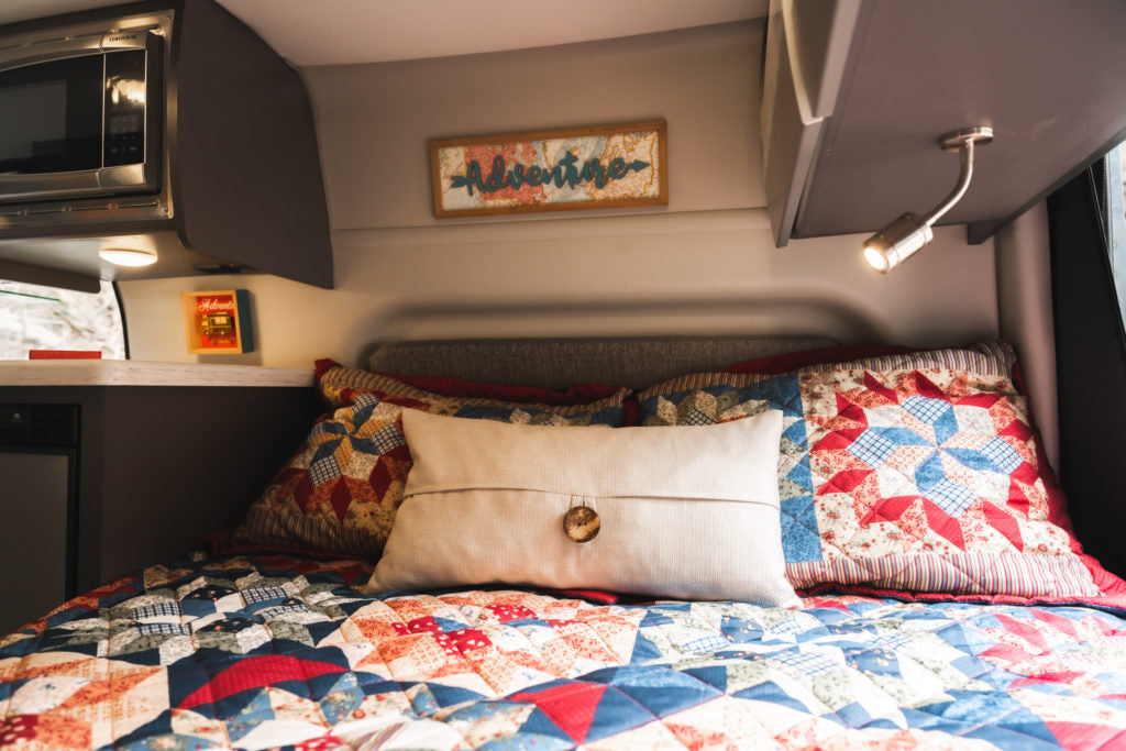 Relax in this cozy RV rental bed! Features a colorful quilt, microwave, and an "Adventure" sign, ideal for road trips.