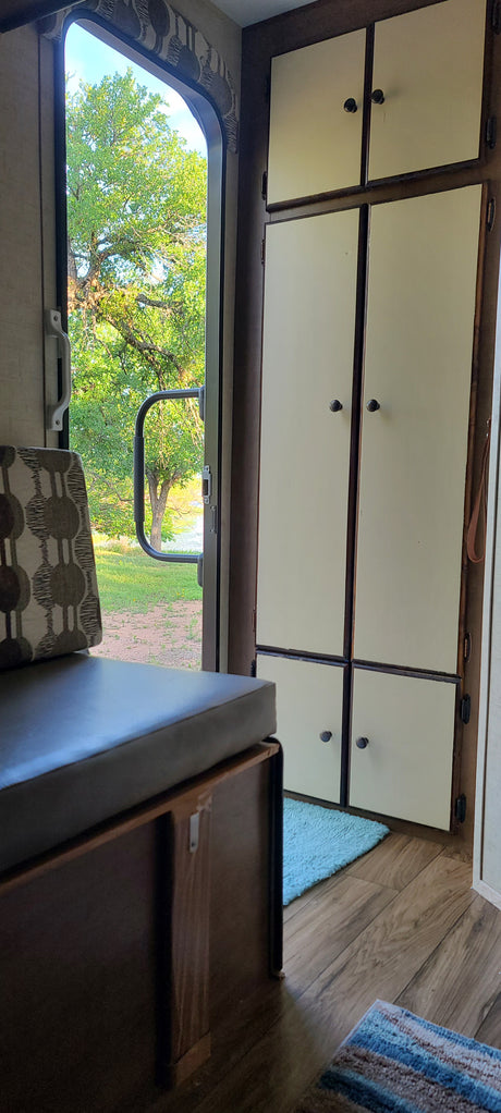 RV rental interior showing bright overhead cabinets, cozy seating, and a wide window view of green trees and water.