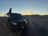A person smiles atop a black RV rental at sunset, on a desert road with mountains. Epic road trip adventure.