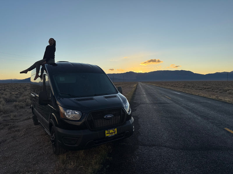 A person smiles atop a black RV rental at sunset, on a desert road with mountains. Epic road trip adventure. — photo 10