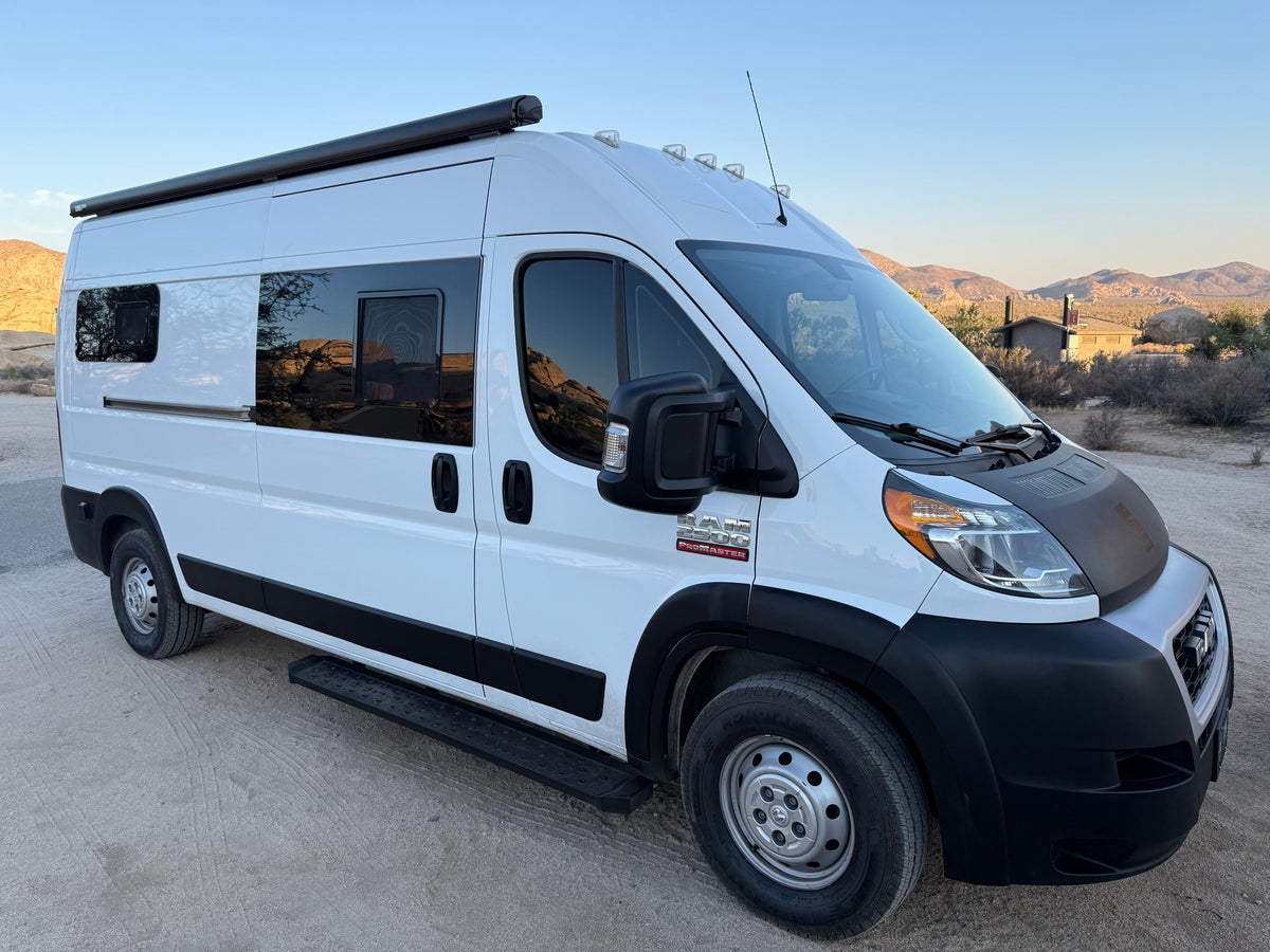 White Ram ProMaster camper van RV rental in a desert setting with mountains in the background, parked on a dirt road.