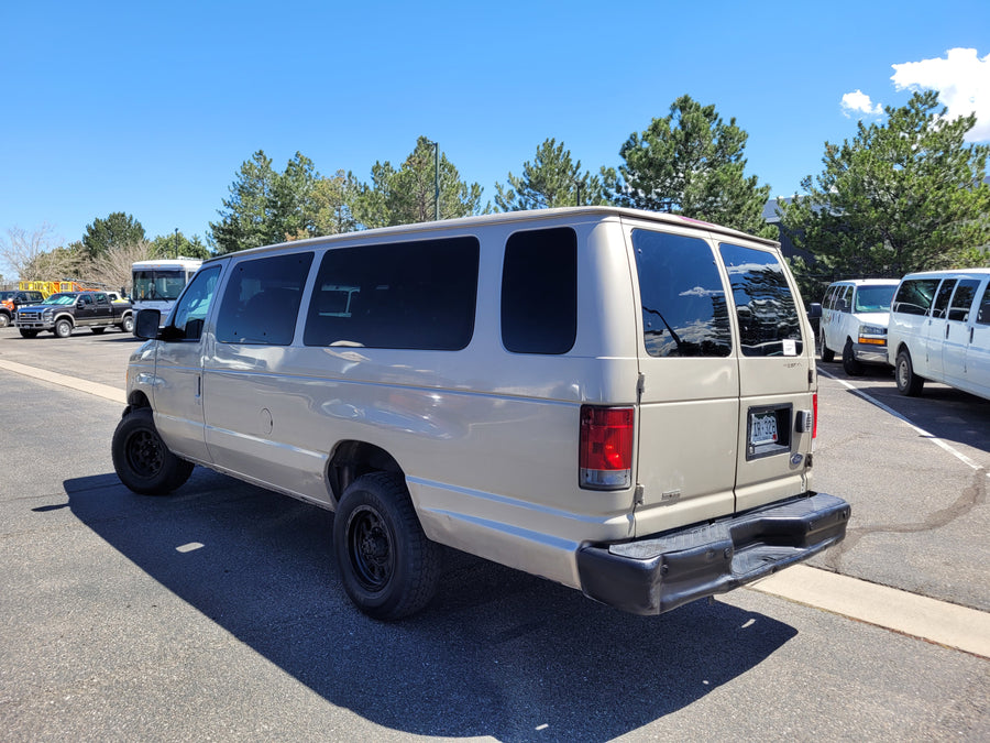 A tan Ford Econoline van, resembling an RV rental, parked on asphalt on a sunny day. Other vans nearby.