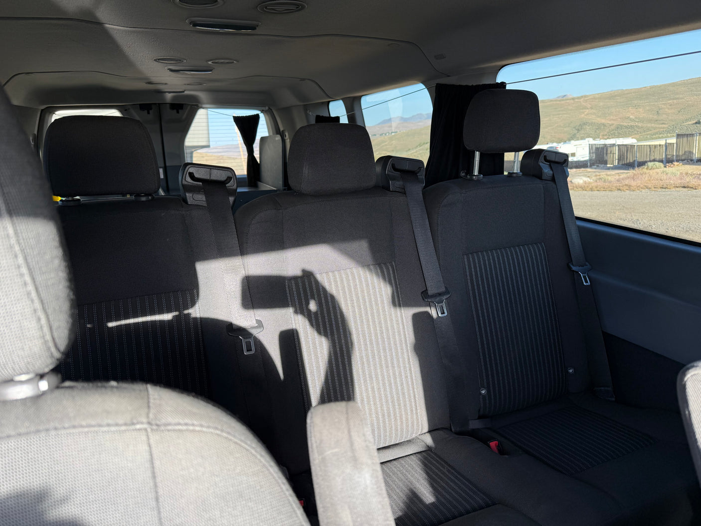 Interior of an **rv rental** van featuring three dark rear seats. Sunlight casts shadows, revealing a landscape with parked RVs outside.