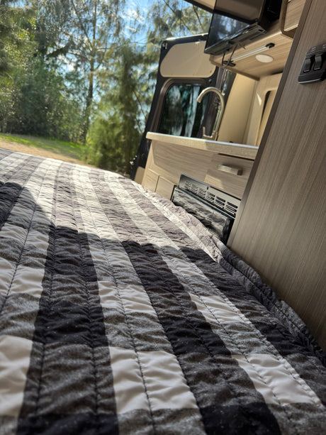 RV rental: inviting bed with black & white striped quilt, mini-kitchen, and serene nature visible through the open door.
