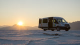 A silver Wandervans RV rental is parked on expansive white salt flats at sunset, doors open with mountains in background.
