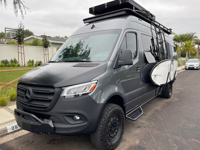 Customized gray Mercedes Sprinter van with off-road tires, roof rack, and surfboard, ready for adventure as an RV rental.