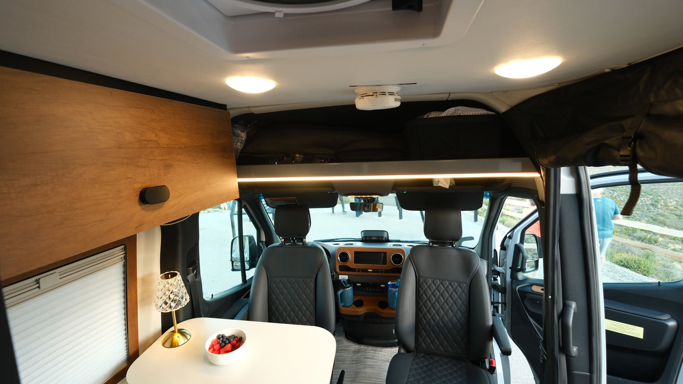 Step inside this cozy RV rental camper van, featuring a driving cabin, dining area with fresh fruit, and ample overhead storage.