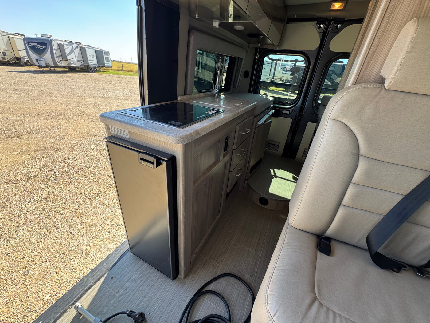 Explore this RV rental's compact interior, featuring a kitchen with a built-in stovetop, sink, and refrigerator, beside a comfortable passenger seat. — photo 10